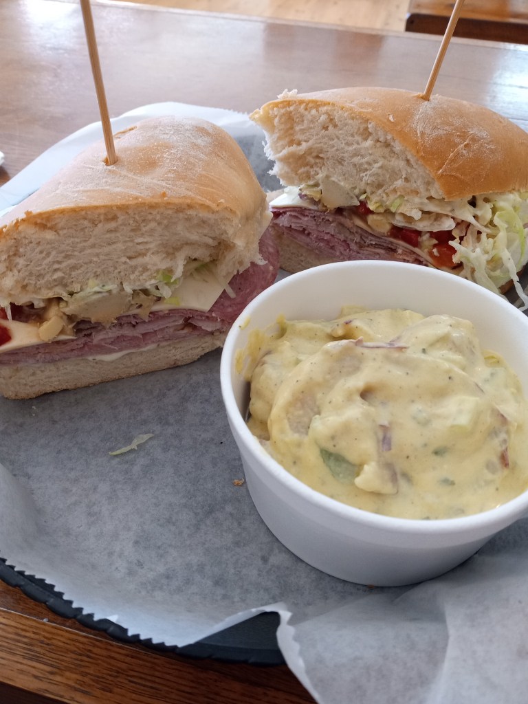 Italian sub cut in half with a side of potato salad in a styrofoam container