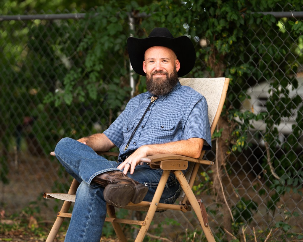 Brandon Bretz sitting in a chair outdoors with a chain link fence and green ivy in the background.