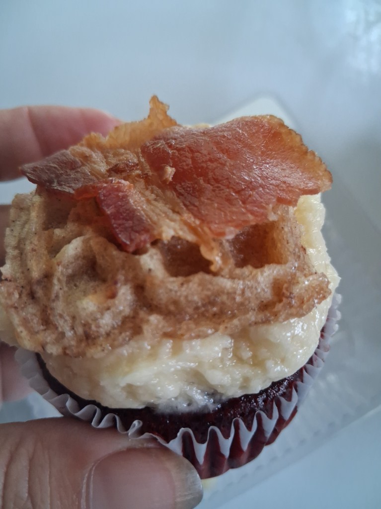 a breakfast cupcake combines red velvet cake and maple flavored frosting, topped with a waffle piece and crispy bacon.