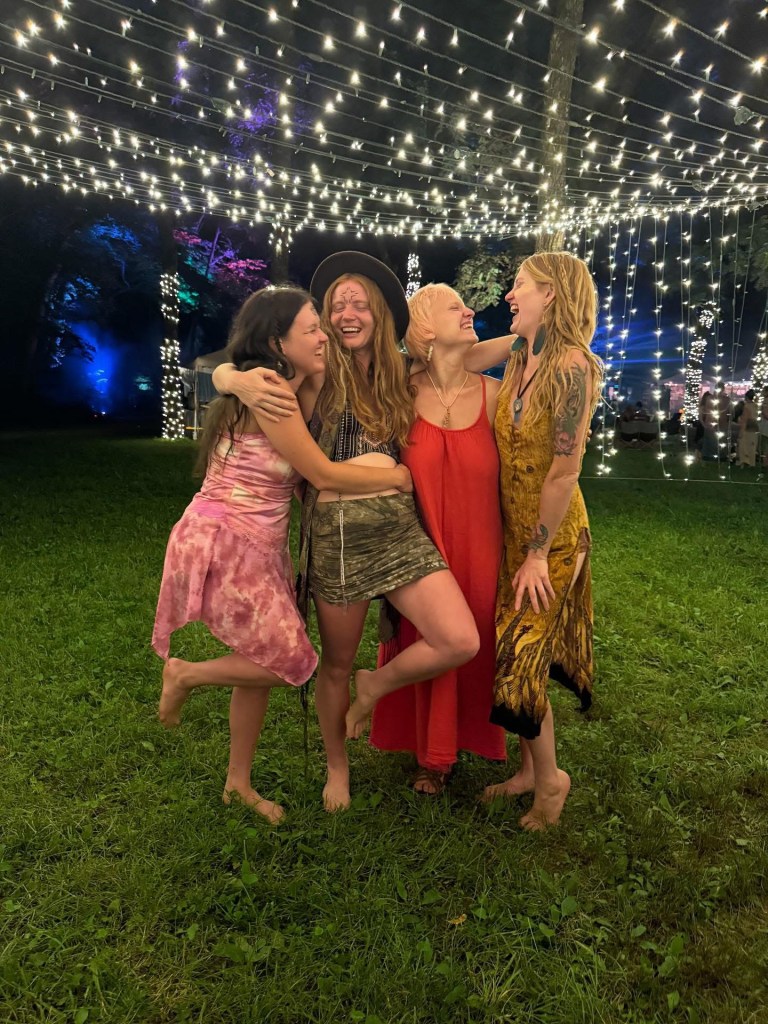 A group of four women posing together outdoors at night, smiling under white string lights