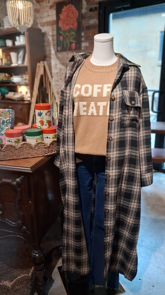 a mannequin wearing a tan sweater that says coffee weather with a long flannel duster over it 